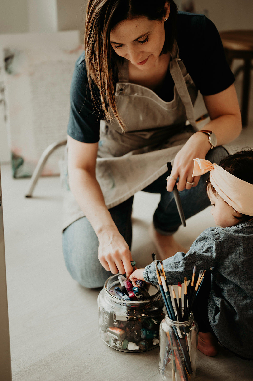 Woman and child sitting on the floor with art supplies, including paintbrushes and a jar of items.